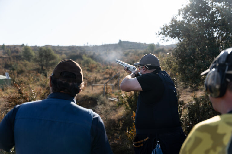 An adult man is shooting during a skeet shooting competition while two more participants are waiting behind. Concept of sport.